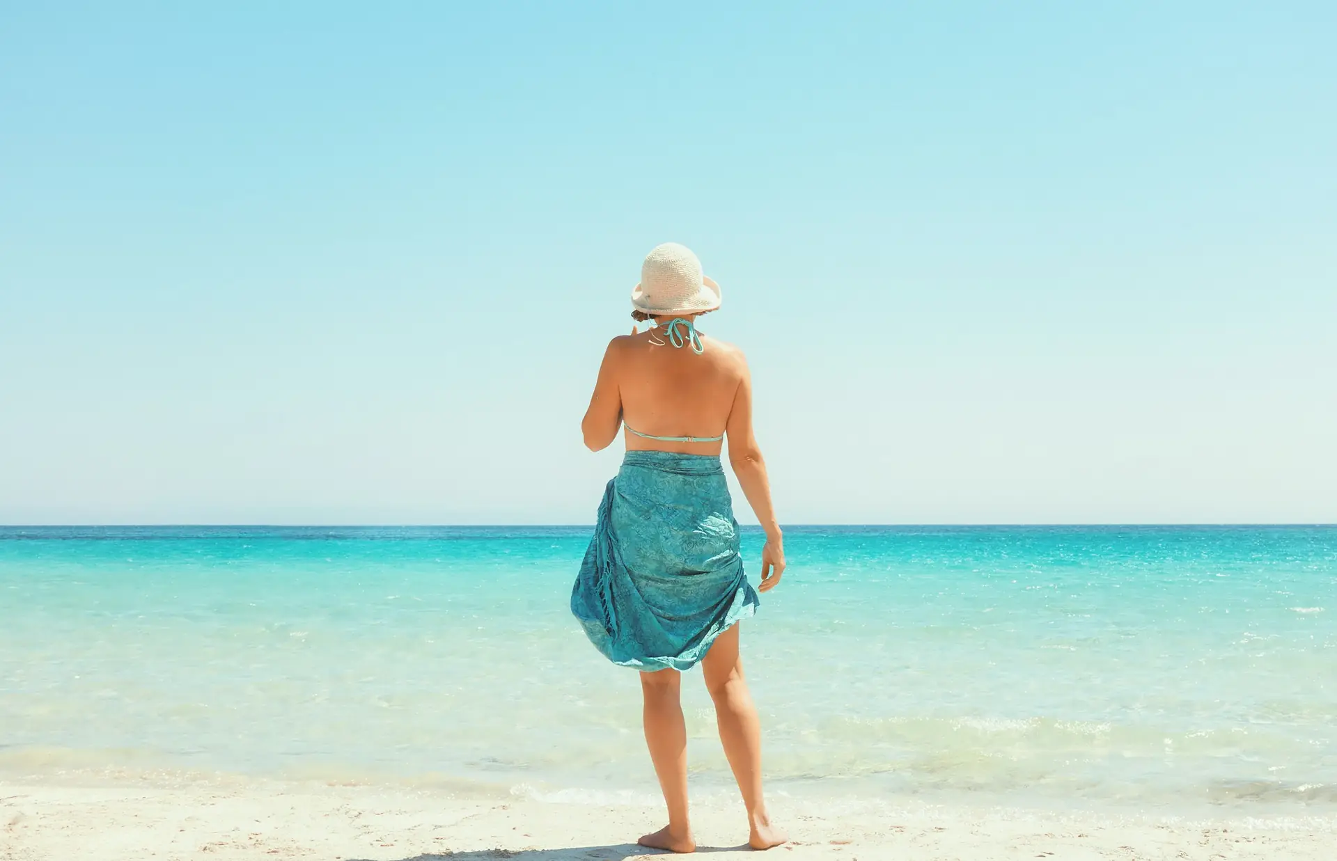 A woman in a sun hat and a blue sarong stands at the edge of a turquoise sea