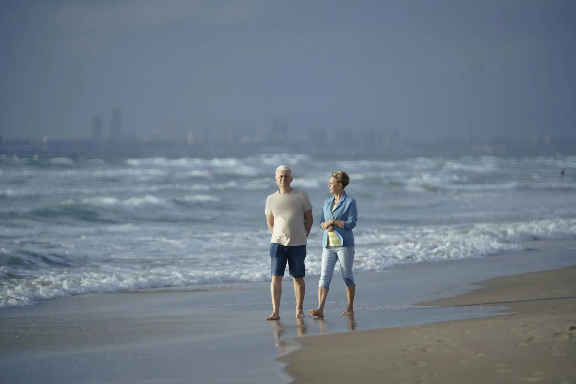 A couple in their fifties stands barefoot at the water's edge on a wide beach